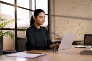 Profissional japonesa organizada trabalhando em um escritório moderno, usando laptop para planejar tarefas e aumentar a produtividade no trabalho.
