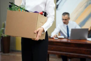 Mulher segurando uma caixa de papelão cheia de pertences pessoais, em frente a um homem sentado em uma mesa de escritório com notebook, sugerindo uma situação de demissão ou desligamento de trabalho