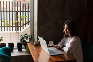 Mulher sentada em uma mesa de madeira em um café, trabalhando em um laptop. Ela usa uma camisa branca, há uma xícara de café ao lado do computador e plantas próximas à janela, com luz natural entrando no ambiente.