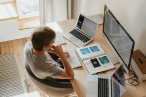 Profissional trabalhando remotamente em estação com múltiplos computadores, representando gestão de ativos de TI da NetEye fora do escritório e monitoramento remoto de dispositivos